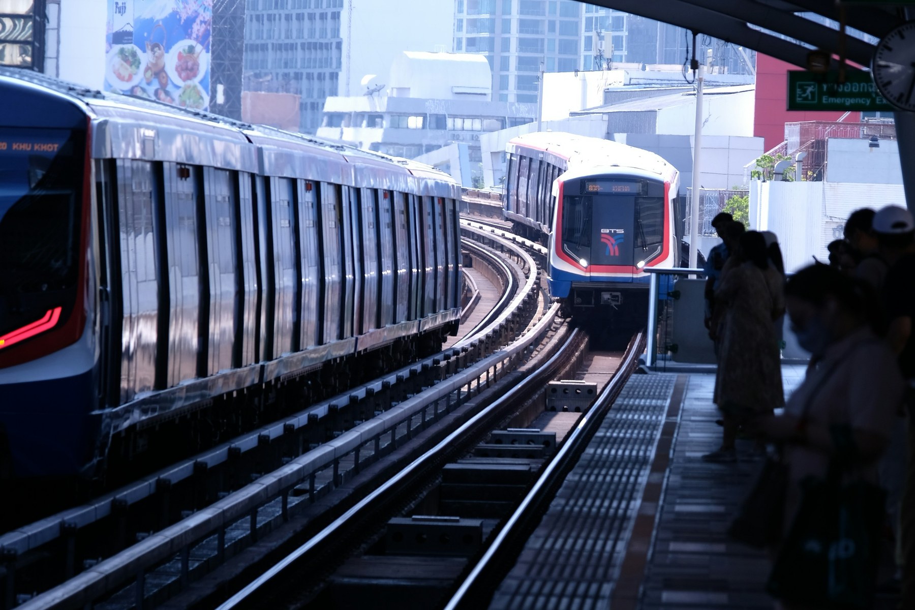 Two BTS Skytrain trains arriving and departing at an elevated Bangkok station.