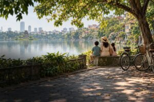 AI-generated editorial illustration of a family seated beneath leafy trees beside a Hanoi-inspired lake, with bicycles and buildings in the background.