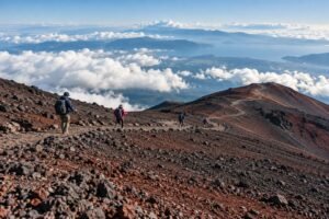 Hikers on a broad volcanic Mount Fuji-inspired trail above clouds in calm summer daylight.
